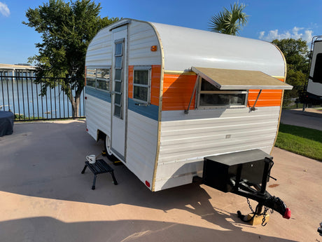 This charming vintage RV rental, a travel trailer with orange and blue accents, sits by a lake, offering a scenic outdoor getaway.