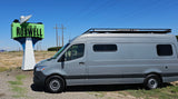 Grey rv rental van with a dog in the driver's seat, parked next to the "Welcome to Roswell" sign under blue sky.