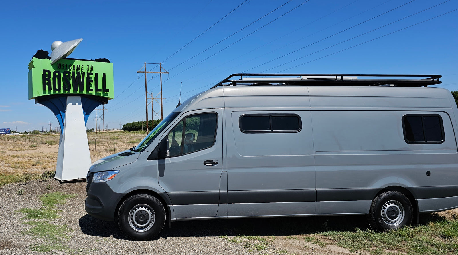 Grey rv rental van with a dog in the driver's seat, parked next to the "Welcome to Roswell" sign under blue sky.