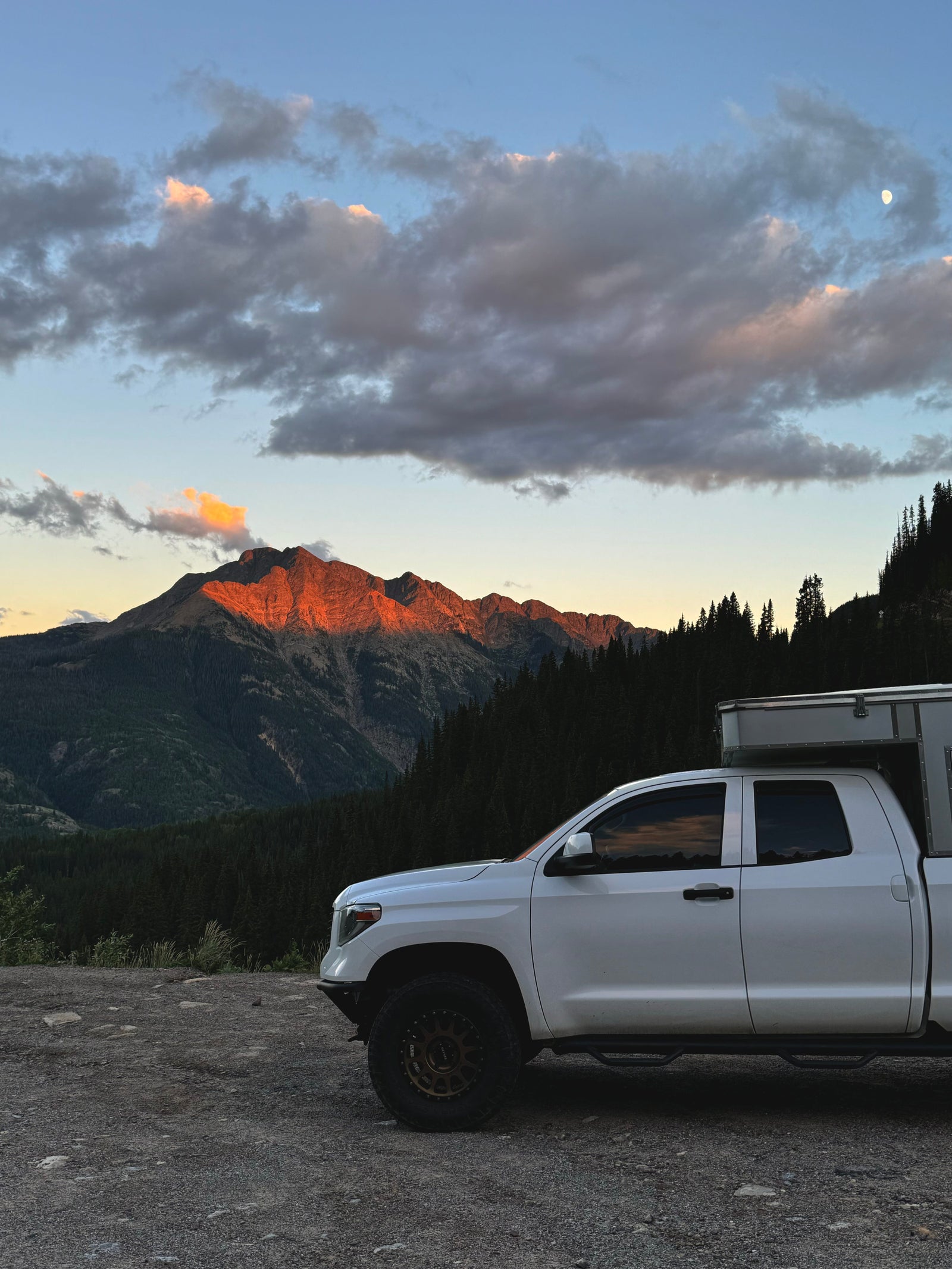 Enjoy a scenic RV rental trip! White truck camper at sunset with a moonlit mountain and forest backdrop.