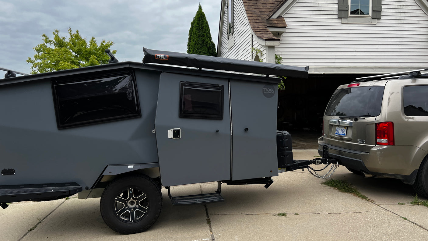 A rugged gray camper trailer, a perfect RV rental option, is hitched to a gold SUV in a driveway, ready for outdoor adventures.