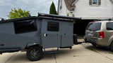A rugged gray camper trailer, a perfect RV rental option, is hitched to a gold SUV in a driveway, ready for outdoor adventures.
