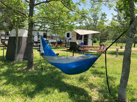 Enjoying a bright RV rental campsite. A blue/gray hammock hangs between trees, with two RVs, a tent, and picnic table nearby.