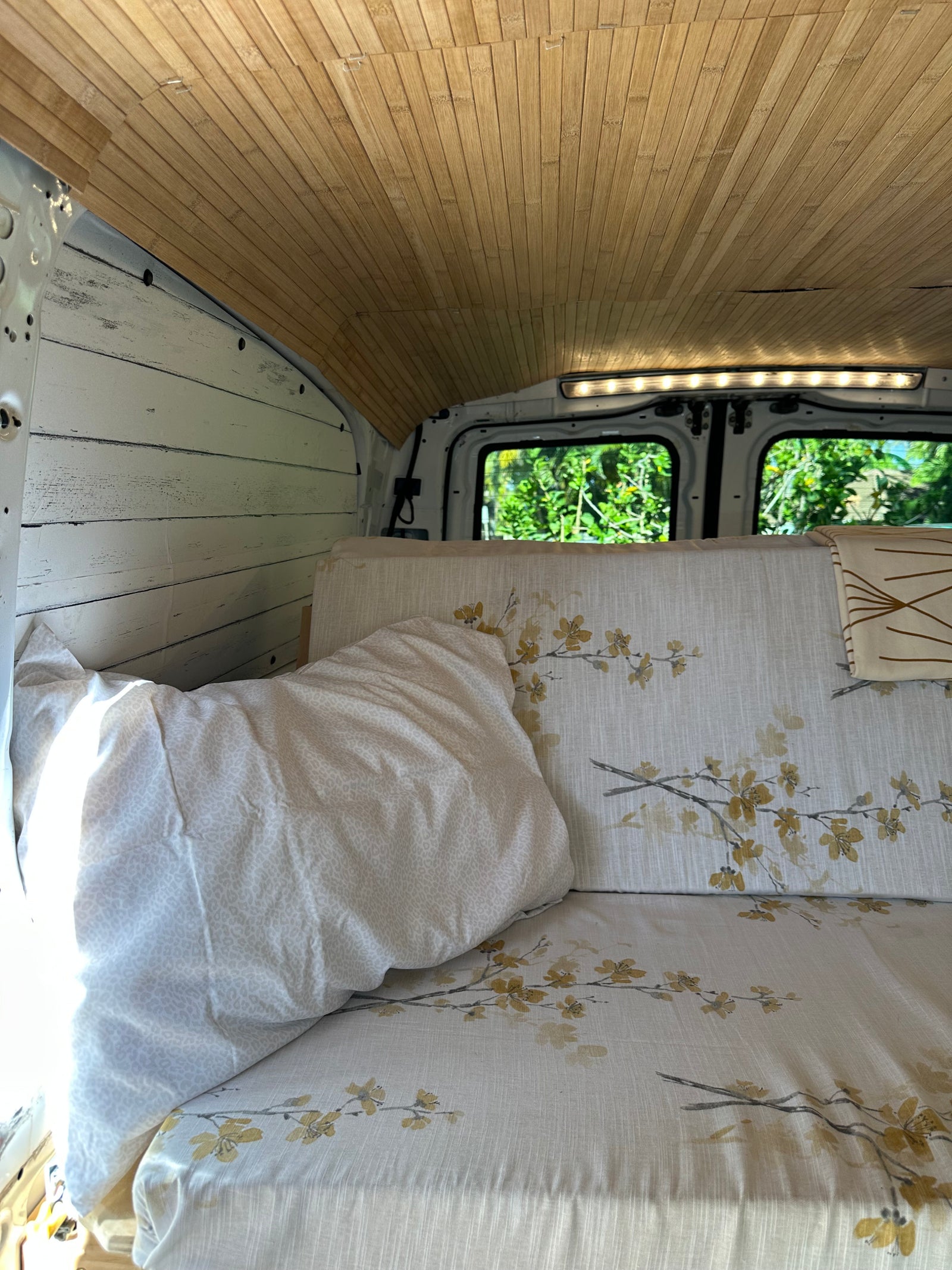Cozy RV rental interior featuring a wood-slat ceiling, white paneling, floral bed, and bright rear window view.