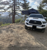 White Toyota Tacoma with rooftop tent set up on a dirt path, overlooking a serene lake. An adventurous RV rental option!