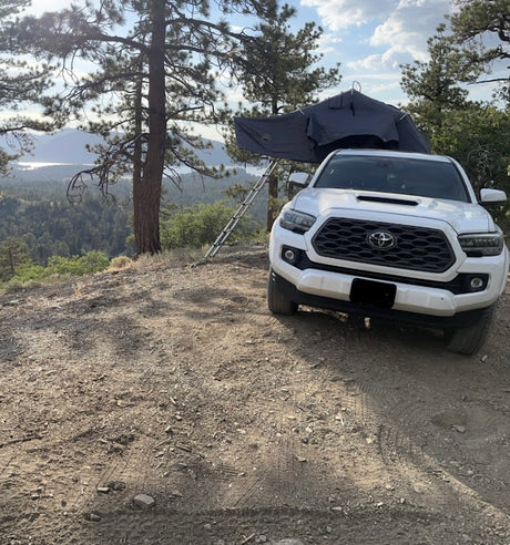 White Toyota Tacoma with rooftop tent set up on a dirt path, overlooking a serene lake. An adventurous RV rental option!