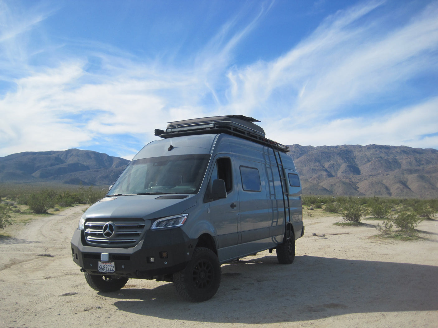 Adventure van RV rental: a grey Mercedes Sprinter in the desert, with a roof rack, ladder, and mountains.