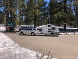 A silver SUV tows a compact travel trailer RV rental through a snowy pine campground, ready for winter camping adventures.