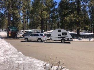 A silver SUV tows a compact travel trailer RV rental through a snowy pine campground, ready for winter camping adventures.