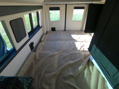 This bright RV rental interior features a spacious, cozy bed with a soft blanket, and large windows revealing green grass outside.