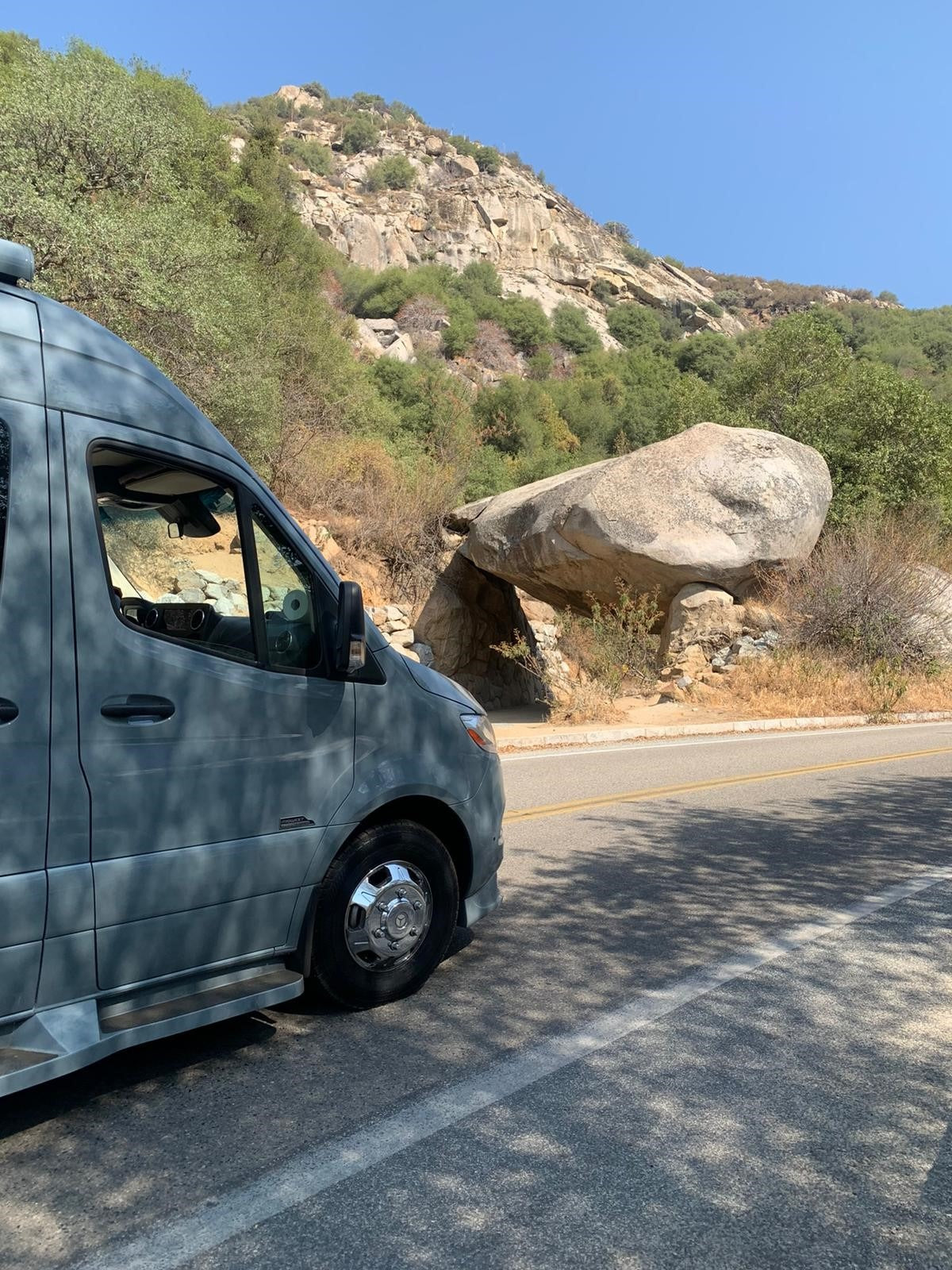 A sleek gray RV rental is parked on a mountain road beside a unique large rock formation under a clear sky.