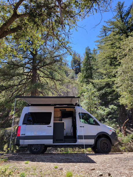 White camper van with open door and extended awning parked on a forest dirt road under blue sky. Perfect for your next RV rental adventure.
