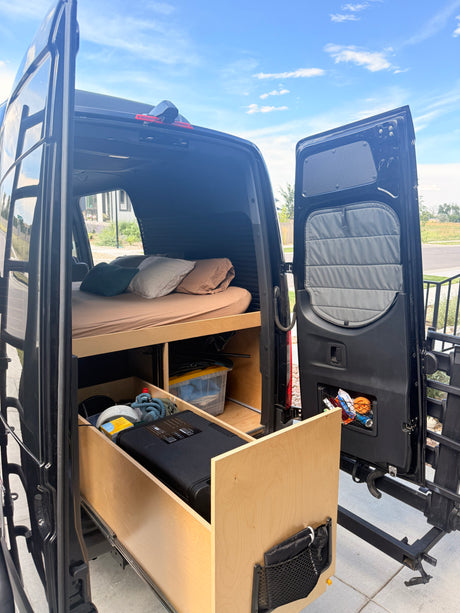 Rear view of an RV rental van's interior, showcasing a bed with pillows and an accessible pull-out storage drawer system.