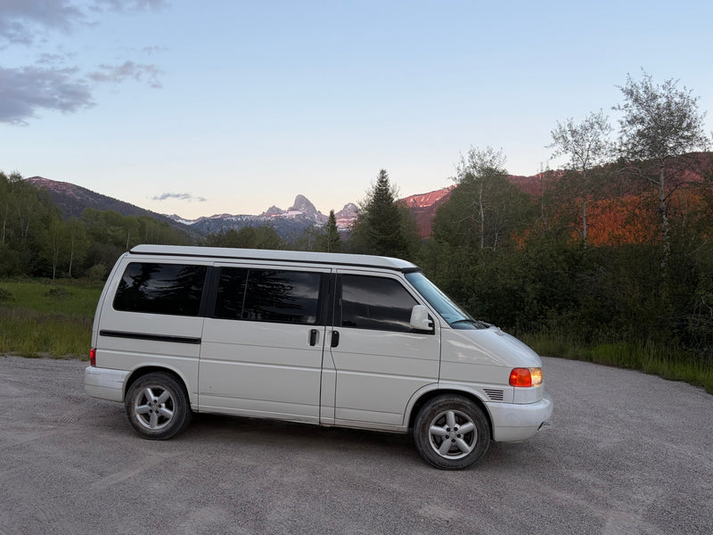 White camper van, likely an RV rental, on a gravel road with the majestic Grand Tetons and forest at dusk. — photo 12
