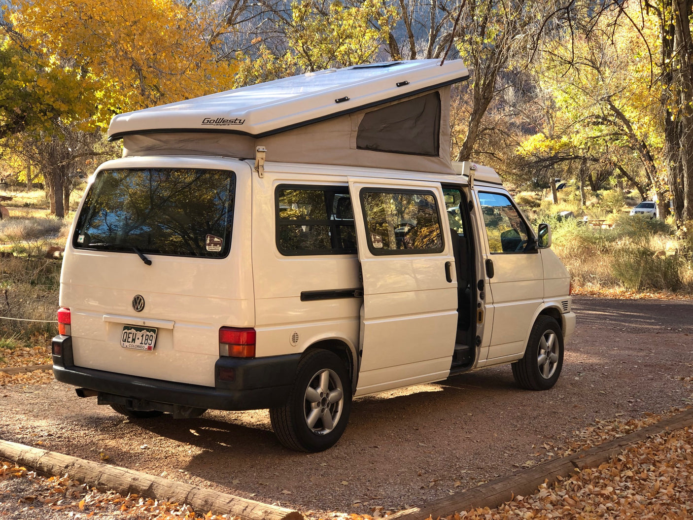 White Volkswagen camper van with pop-top up, side door open, parked at an autumn campsite. Ideal rv rental for fall trips!