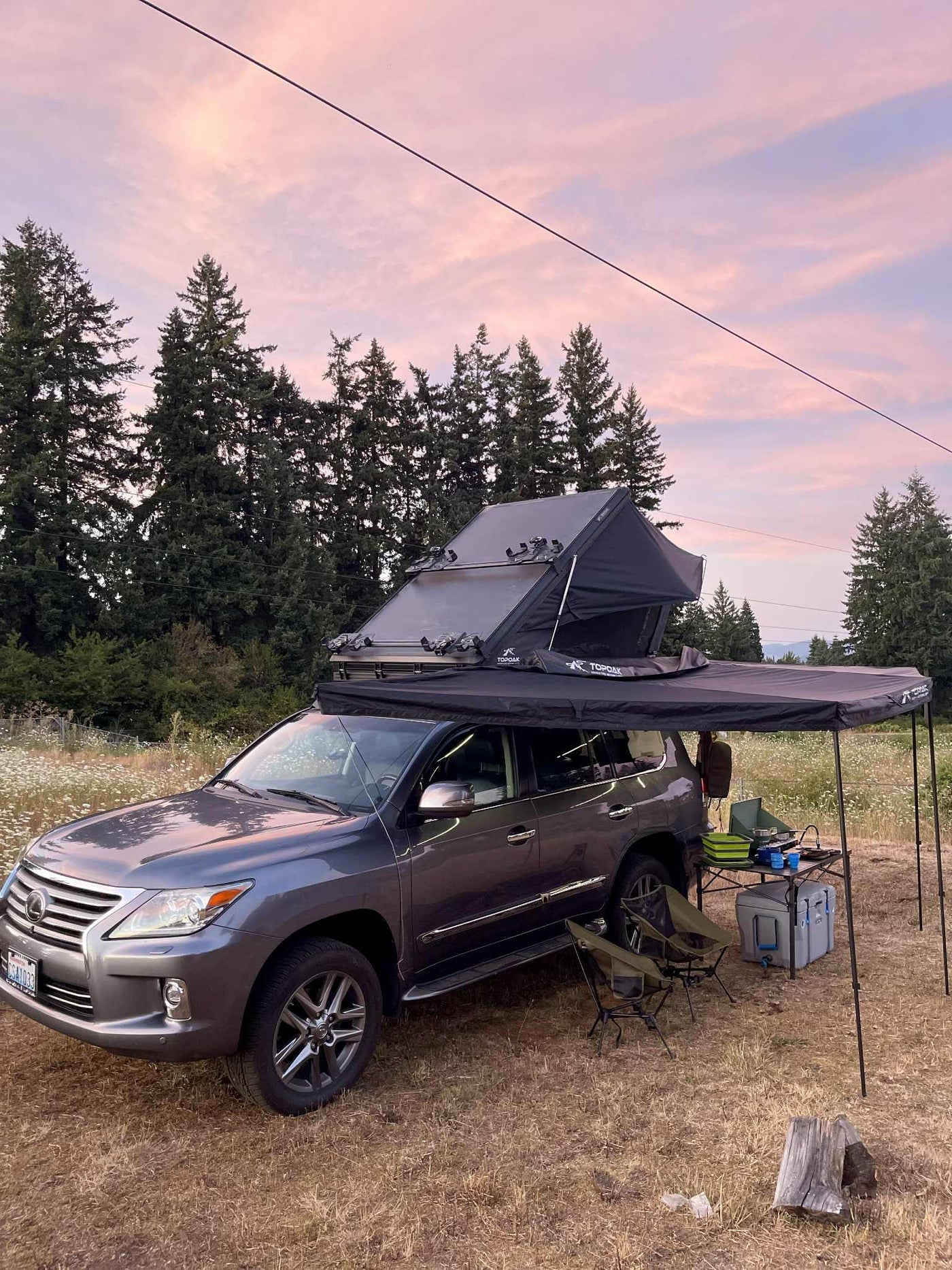 Grey Lexus SUV with rooftop tent and awning. This RV rental alternative is set up for camping at sunset amidst tall trees. — photo 8