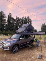 Grey Lexus SUV with rooftop tent and awning. This RV rental alternative is set up for camping at sunset amidst tall trees.