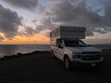 Coastal rv rental with a view: White truck camper parked by the ocean at sunset, showcasing a serene outdoor adventure.