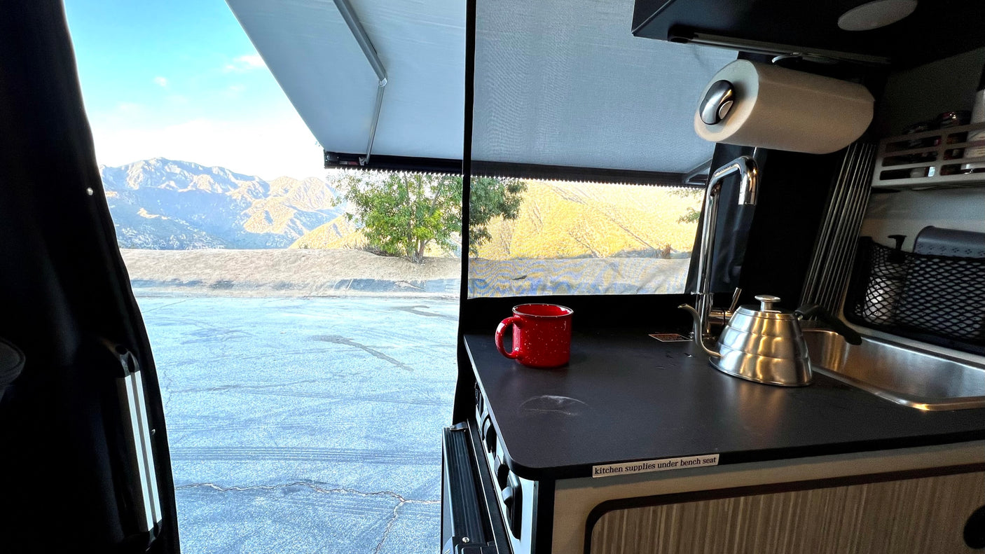 RV rental camper interior: kitchen counter with red mug & kettle. Outside, a mountain view under an open awning.