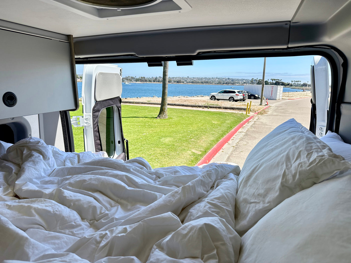 Comfy white bed inside an rv rental, featuring a wide-open view of a serene bay and green park.