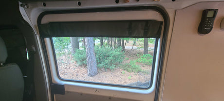 RV rental view from inside: a window with a shade looks out to pine trees & a remote on the wall.