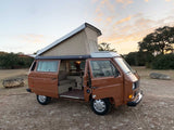 Brown VW Vanagon camper pop-top RV rental at sunset. Side door open revealing interior, parked on gravel in nature.