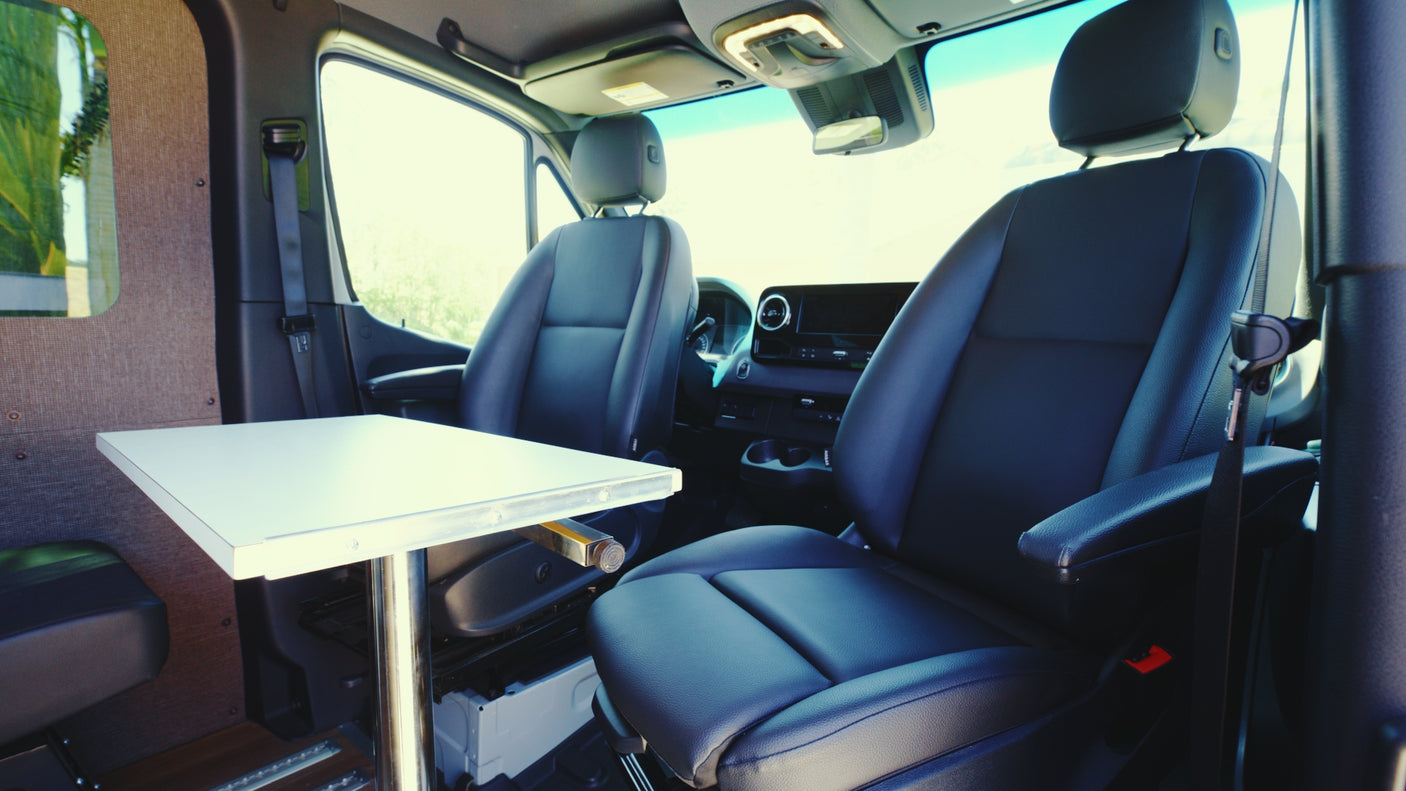 RV rental interior: two black front seats, a white table with chrome leg, and dashboard details visible, shot from the rear cabin.