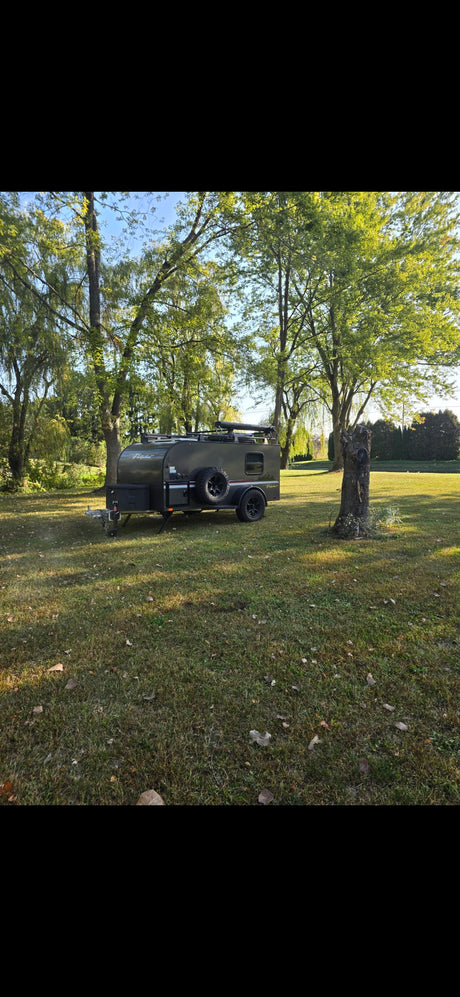 This dark green, rugged teardrop camper, ideal for an RV rental, is parked on a sunny, grassy lawn among trees.