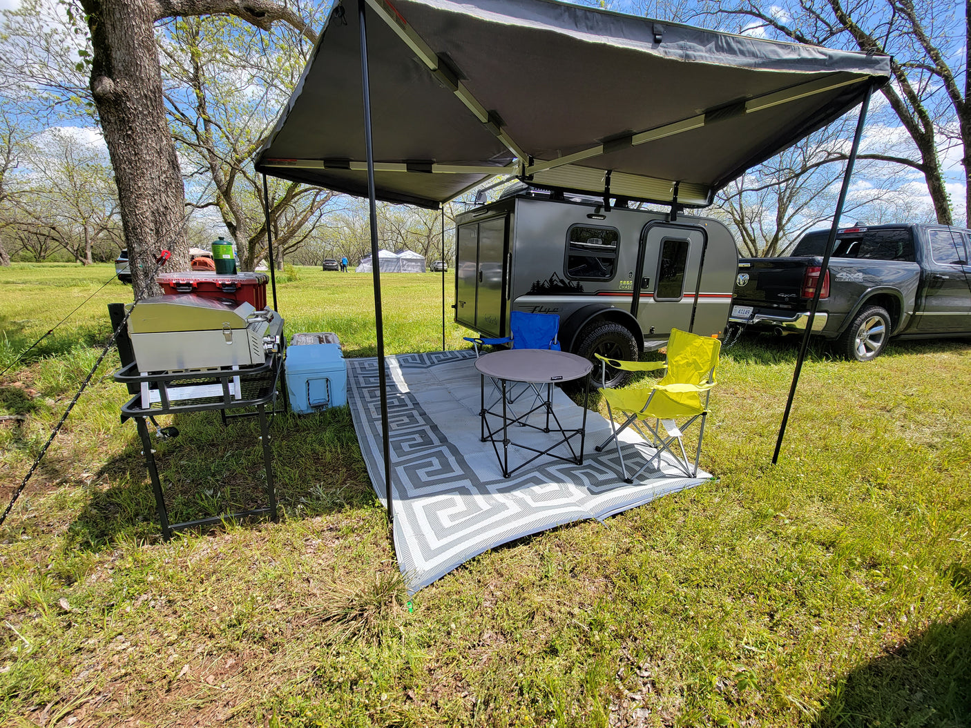 Perfect RV rental setup: small travel trailer with an extended awning covering a picnic area with table, chairs, & grill.