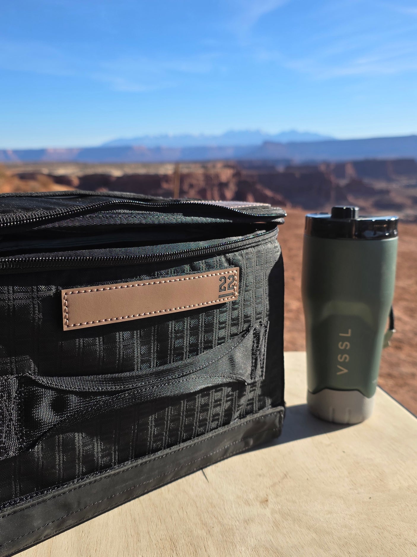 Road trip views! This RV rental stop features a black bag and VSSL thermos on a wooden surface, overlooking a stunning canyon.