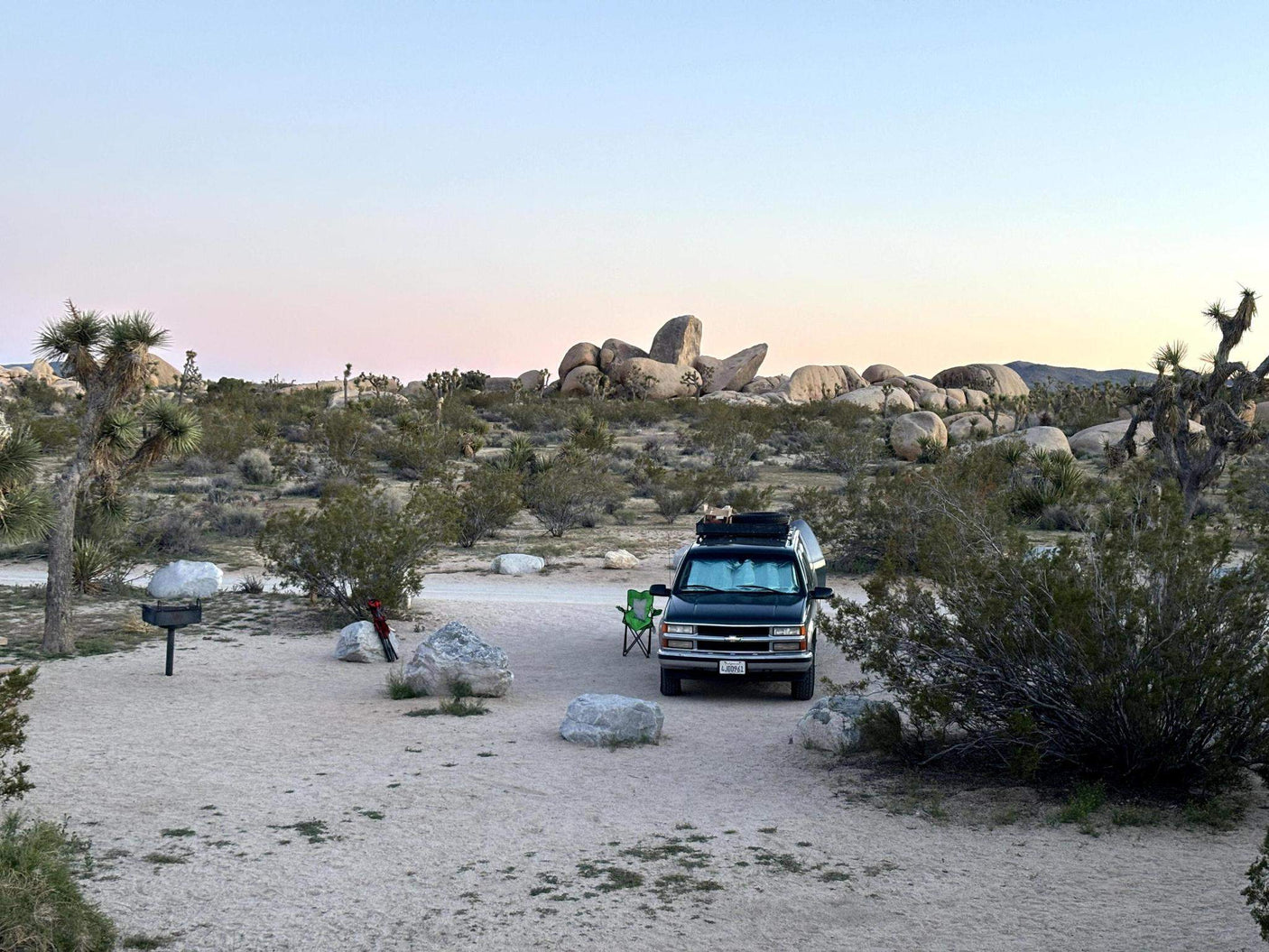 RV rental camping in Joshua Tree National Park at dusk. SUV on a sandy campsite with a grill, rocks, and iconic trees.