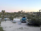 RV rental camping in Joshua Tree National Park at dusk. SUV on a sandy campsite with a grill, rocks, and iconic trees.
