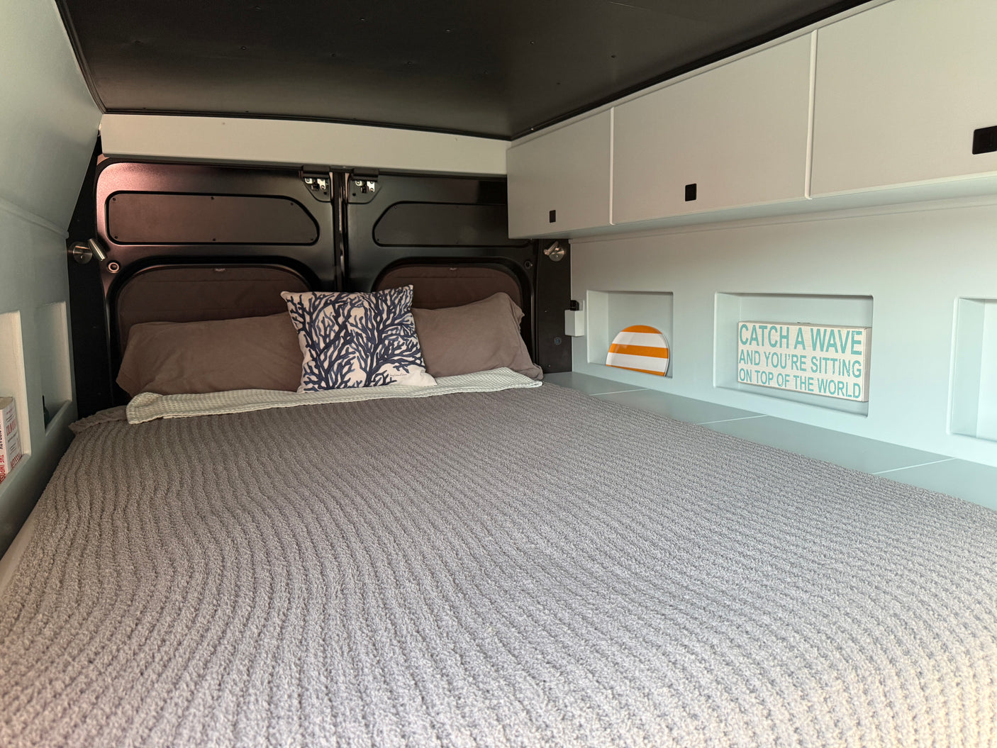An RV rental bedroom with a large made bed, grey bedding, blue coral pillow, and smart storage for a cozy vibe.