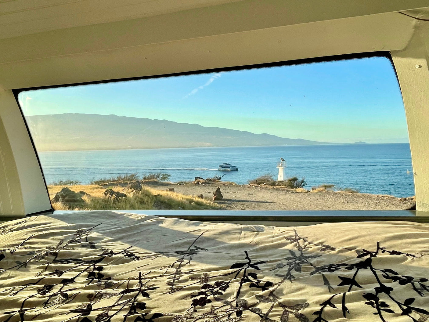 An rv rental bed frames a window view of a sunny coast: mountains, ocean with a boat, and a lighthouse on the rocky shore.