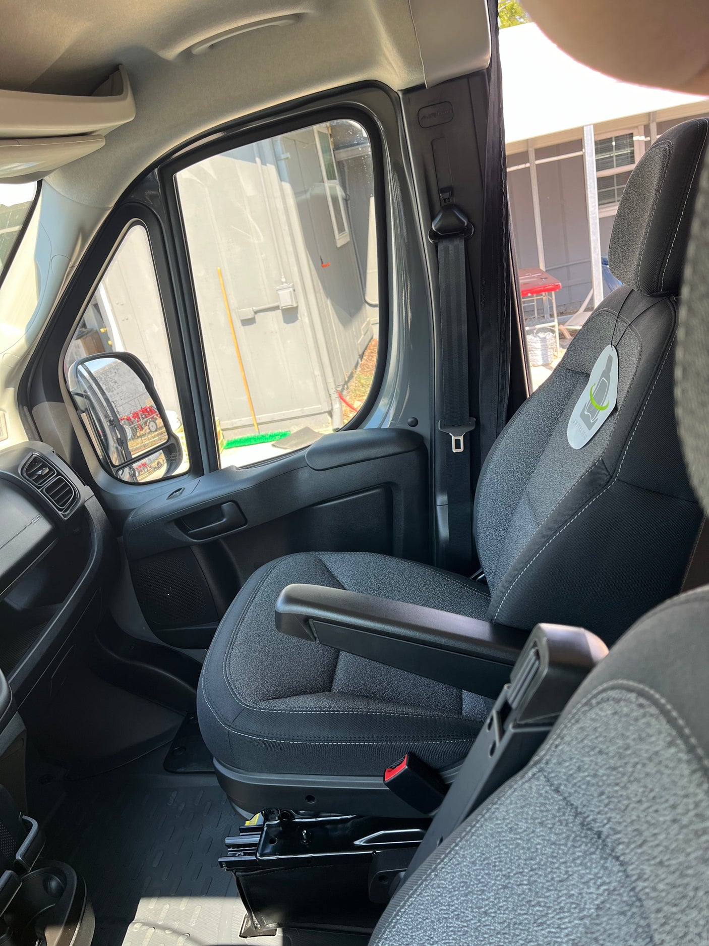 Inside an RV rental, showing the front passenger seat with armrest, dashboard, and side mirror view of outdoor structures.