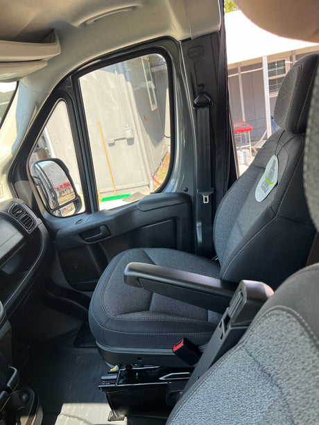 Inside an RV rental, showing the front passenger seat with armrest, dashboard, and side mirror view of outdoor structures.