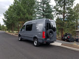 This gray Sprinter van, perfect for rv rental, is set up for adventure with off-road tires, roof rack, and rear ladder.