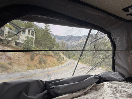 Scenic mountain road & house view from an RV rental's mesh window. Cozy interior with blankets, ideal for road trip travel.