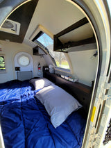 Cozy rv rental camper interior, featuring a bed with a blue comforter and light pillows, overhead storage, and large windows.