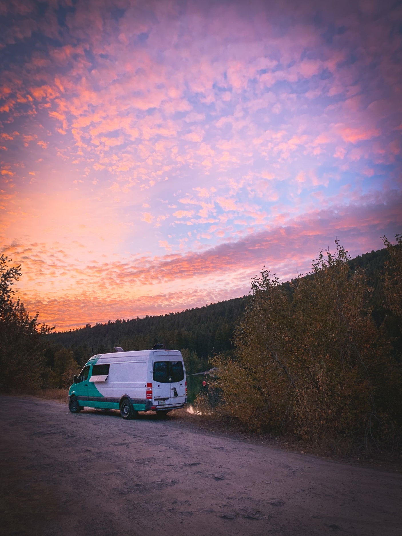 RV rental adventure: a teal and white camper van parked lakeside on a dirt road under a stunning pink and orange sunset.