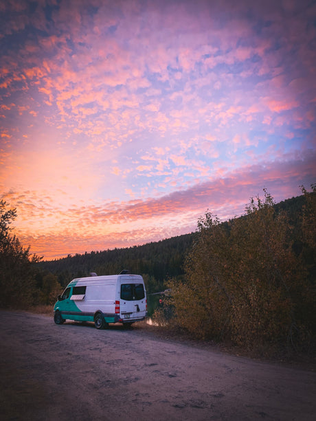 RV rental adventure: a teal and white camper van parked lakeside on a dirt road under a stunning pink and orange sunset.
