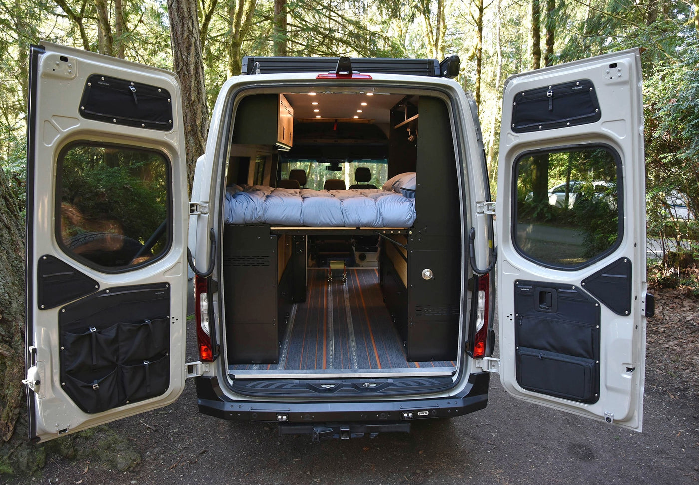 Open rear doors of an RV rental camper van reveal a cozy interior with a raised bed, storage, and patterned wooden floor. — photo 11