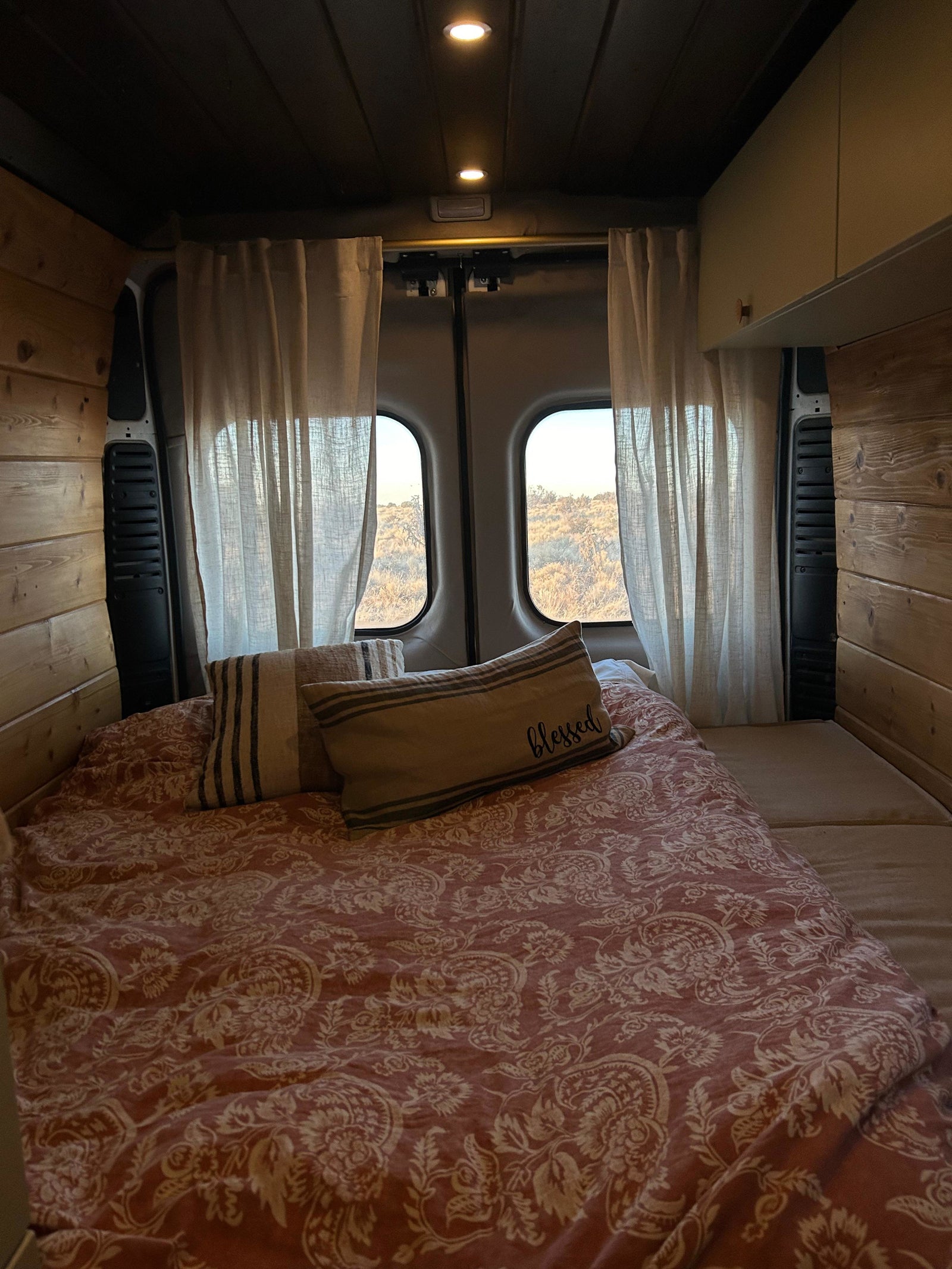 Cozy RV rental bedroom with a red patterned bed, wooden walls, warm lighting, and a scenic view of a dry landscape from the back windows.