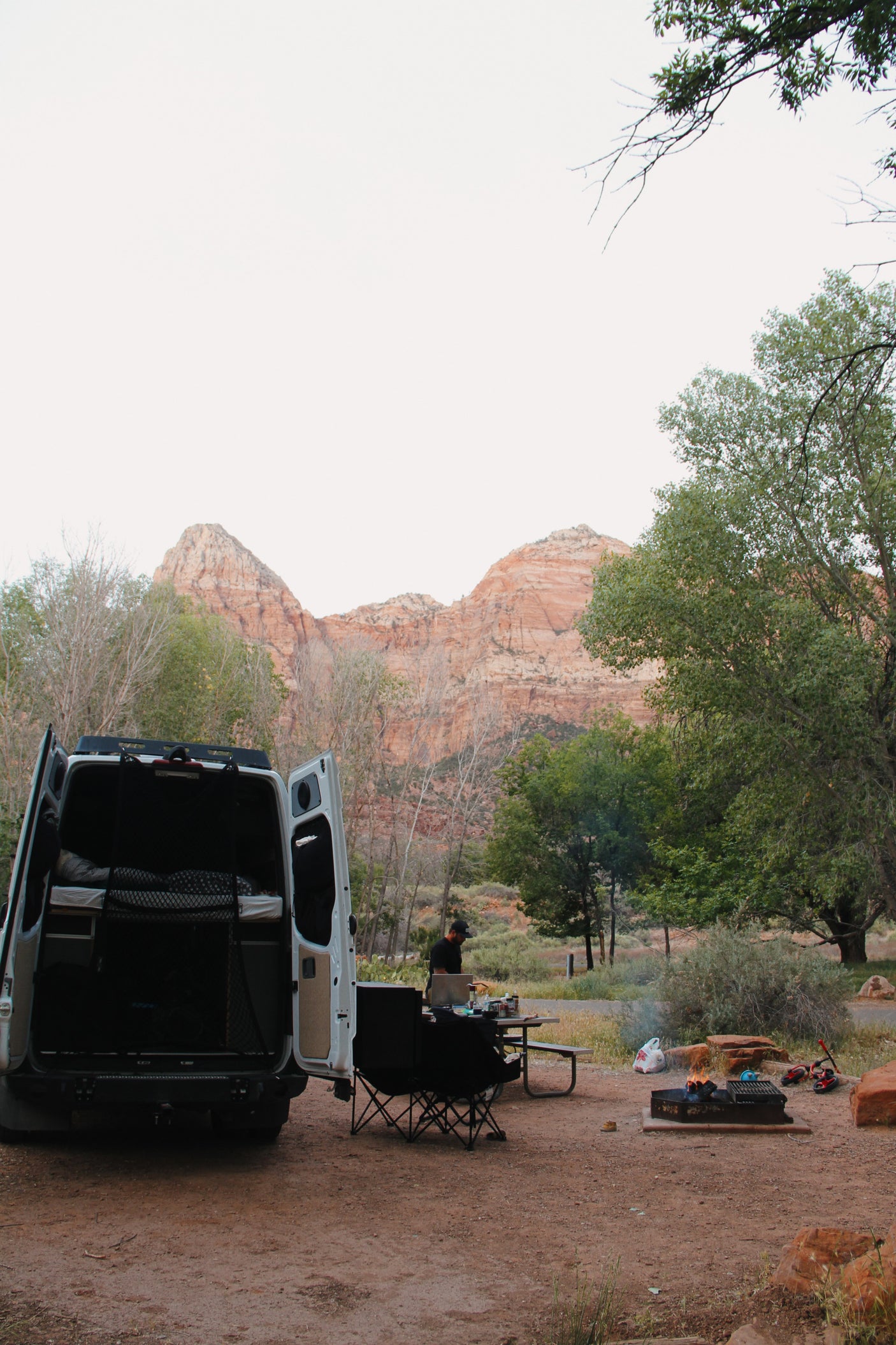 A white RV rental camper van parked at a dirt campsite with a person by a table, chairs, fire pit, and mountains.