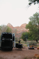A white RV rental camper van parked at a dirt campsite with a person by a table, chairs, fire pit, and mountains.