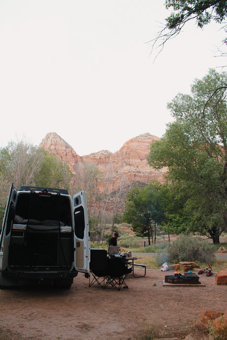 A white RV rental camper van parked at a dirt campsite with a person by a table, chairs, fire pit, and mountains.