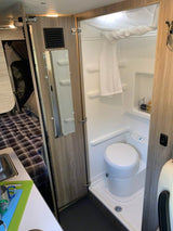 Compact RV rental interior featuring a full bathroom with shower and toilet, alongside a glimpse of the bed and kitchen area.
