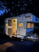 Charming vintage rv rental at night, parked under dark trees. Its open door reveals a warm, lit interior.