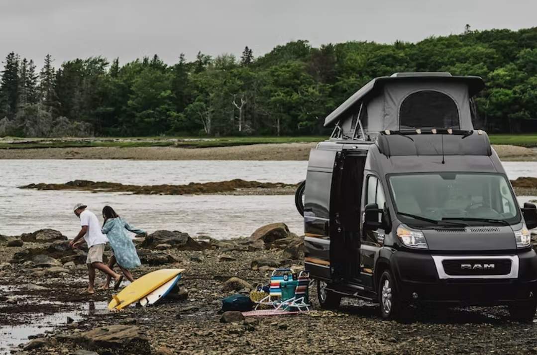 RV rental camper van with pop-top on a rocky shore. A couple walks towards the water, their surfboard nearby for outdoor fun.
