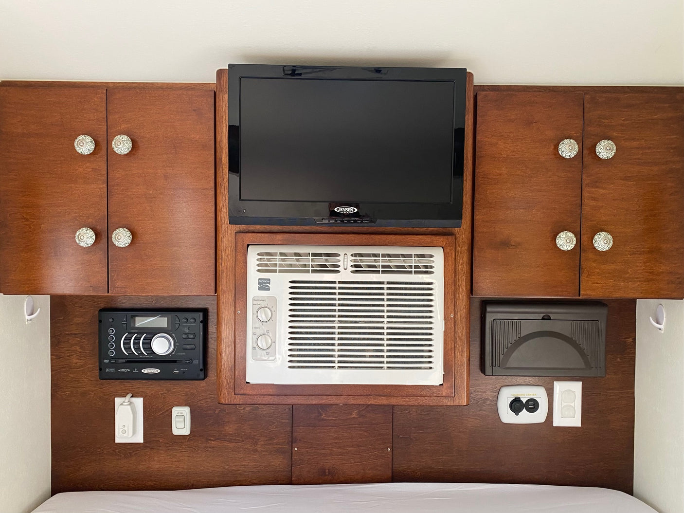 Inside an RV rental: overhead cabinets flank a TV and AC unit, with a stereo, charging center, and outlets below, above a bed.